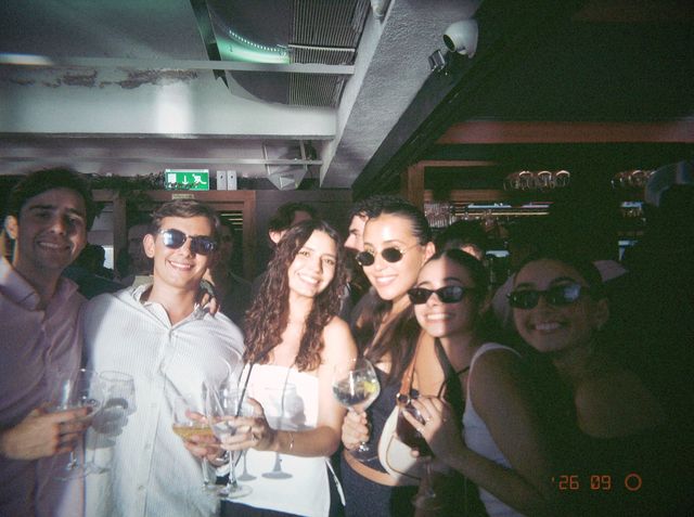 Dos chicos y cuatro chicas, varios con gafas de sol, sonríen apiñados junto a la barra con copas de balón.