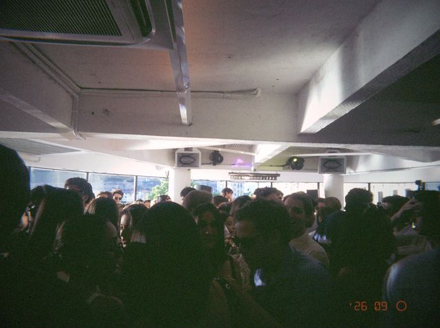 La sala se llena de gente a contraluz, con altavoces en el techo y ventanales con luz de día al fondo.