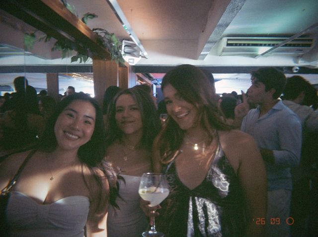 Tres chicas posan sonrientes entre la gente y la de la derecha sostiene una copa de balón con pajita.