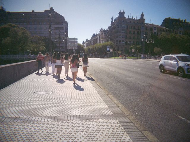 Un grupo de chicas camina de espaldas por una acera soleada de baldosas hexagonales, con edificios señoriales al fondo.