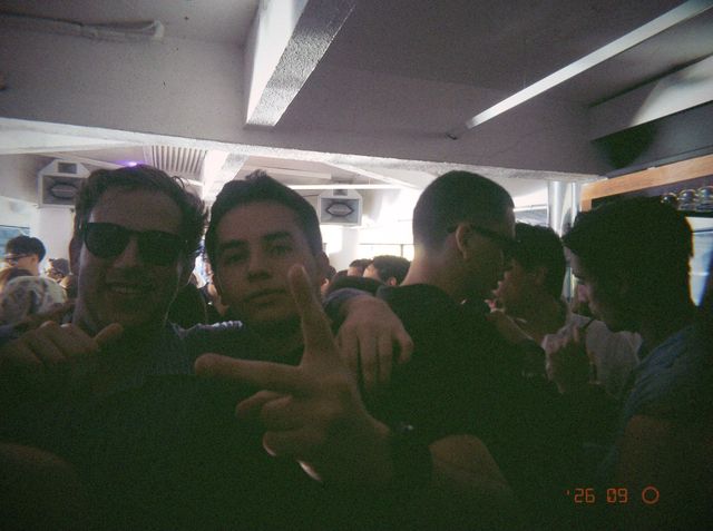 Dos chicos posan en la sala llena, uno con gafas de sol y otro con el índice y el pulgar extendidos hacia la cámara.