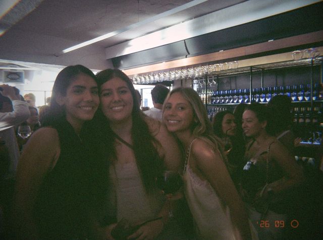 Tres chicas juntan las caras y sonríen en la barra, y una de ellas sostiene una copa de vino tinto.