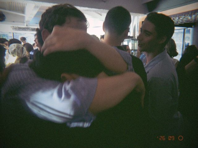 Dos amigos se funden en un abrazo en medio de la sala mientras otros charlan detrás a la luz del día.