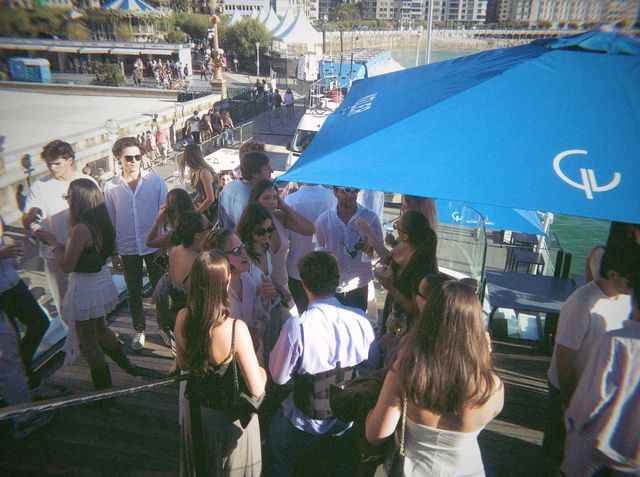 Un grupo de amigos charla al sol en la terraza bajo una sombrilla azul, con el paseo y un tiovivo al fondo.
