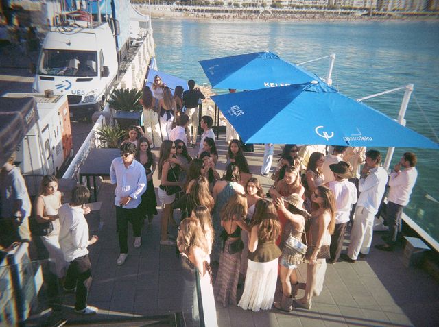 Un grupo numeroso charla en la terraza bajo sombrillas azules de Keler, con la bahía y la playa al fondo.