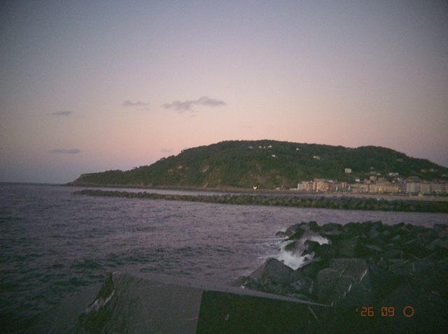 El mar rompe contra las rocas del espigón frente a un monte verde con casas, bajo un cielo rosado.
