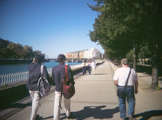 Dos chicos con mochilas caminan por un paseo arbolado junto al río, con un puente al fondo.