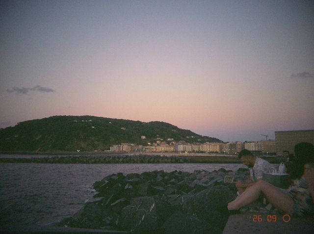 Un chico y una chica se sientan en el espigón de rocas al atardecer, frente a la playa y el monte bajo el cielo rosa.