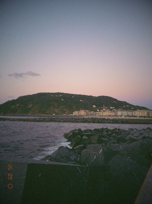 El mar rompe contra las rocas del espigón al atardecer, con un monte verde y una hilera de edificios al fondo.