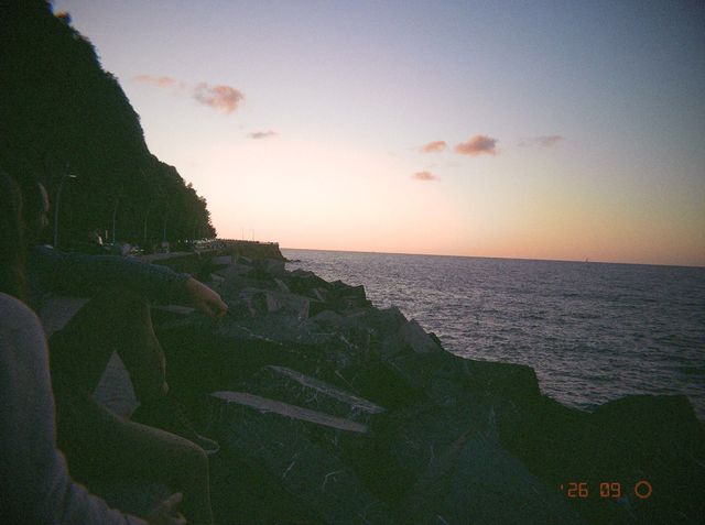 Varias personas se sientan en las rocas junto al mar mientras el cielo se tiñe de rosa en el horizonte.