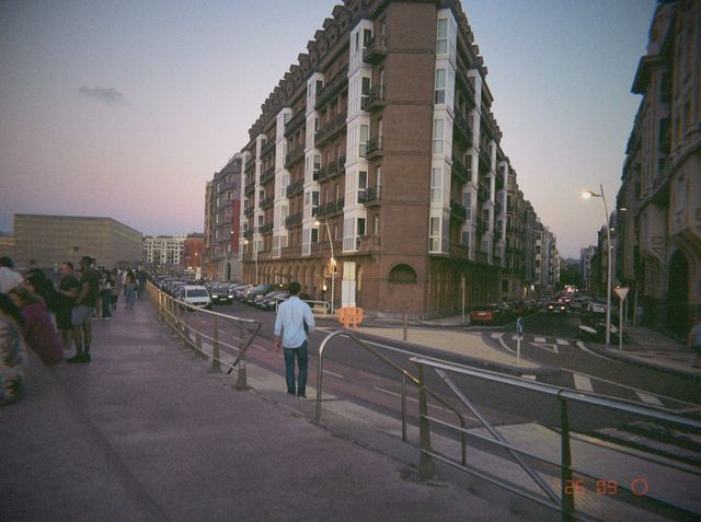 Un chico con camisa azul claro camina junto a la barandilla hacia un gran edificio de miradores blancos al anochecer.