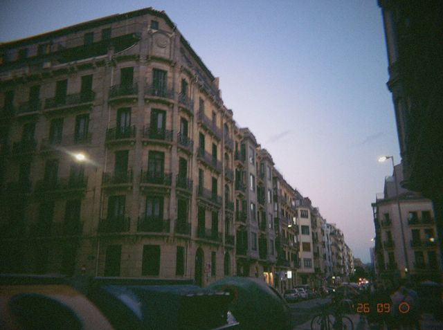Una calle de edificios señoriales con balcones al anochecer, con las farolas ya encendidas y contenedores delante.