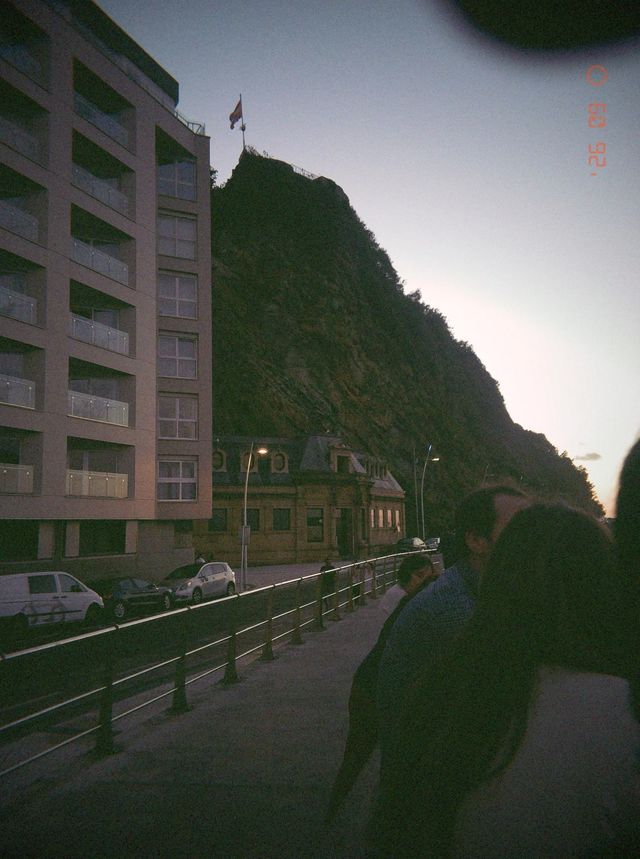 Una pareja se abraza en la acera al anochecer, bajo un monte rocoso coronado por una bandera.