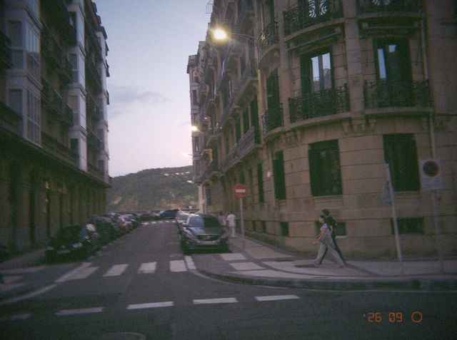Dos personas caminan por una calle de edificios de piedra con balcones de hierro, con las farolas ya encendidas.