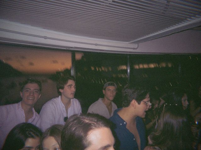 Tres chicos con camisa, uno con gorra del revés, asoman entre la gente ante ventanas con el cielo del ocaso.