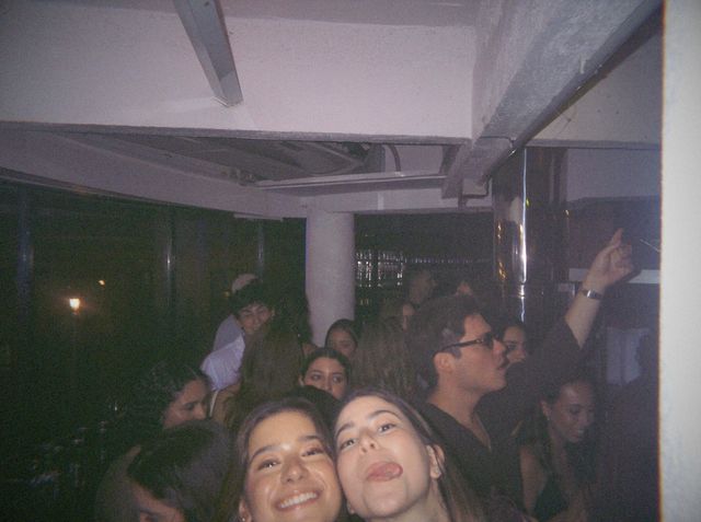 Dos amigas se asoman a la cámara, una sonriendo y otra sacando la lengua, con la sala llena de gente detrás.