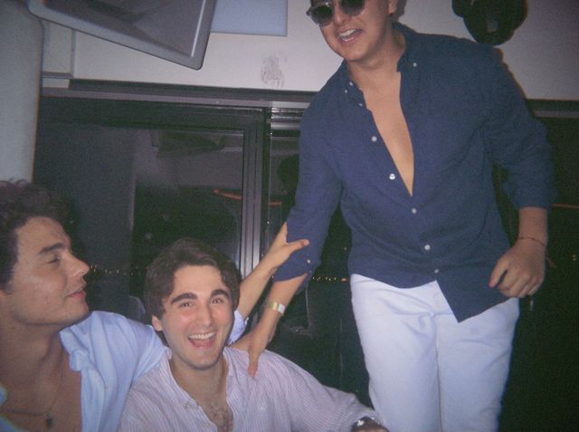 Un chico sentado ríe a carcajadas entre un amigo que lo mira y otro de pie con gafas de sol y camisa azul marino.