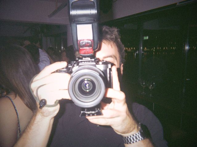 Un chico con reloj plateado apunta con una cámara de fotos grande, con luces lejanas tras los ventanales oscuros.