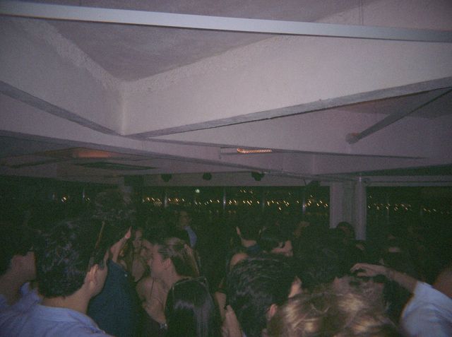 La gente baila apretada en la pista bajo el techo blanco, con las luces de la ciudad tras las ventanas.