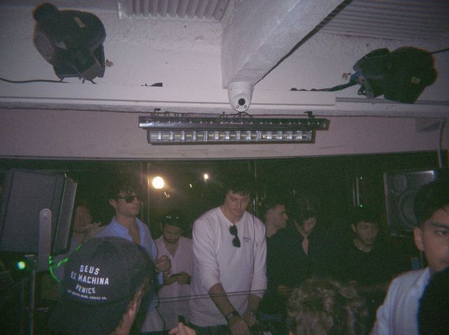 El DJ pincha con camiseta blanca y unas gafas de sol colgadas del cuello, rodeado de chicos en la cabina.