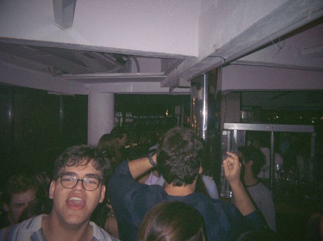 Un chico con gafas canta con la boca abierta mientras detrás otro levanta la mano cerca de la barra.