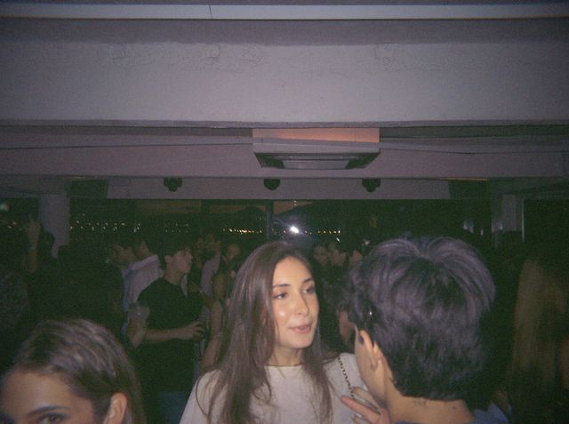 Una chica de pelo largo charla con alguien de pelo corto en la sala llena, con luces lejanas tras los ventanales.