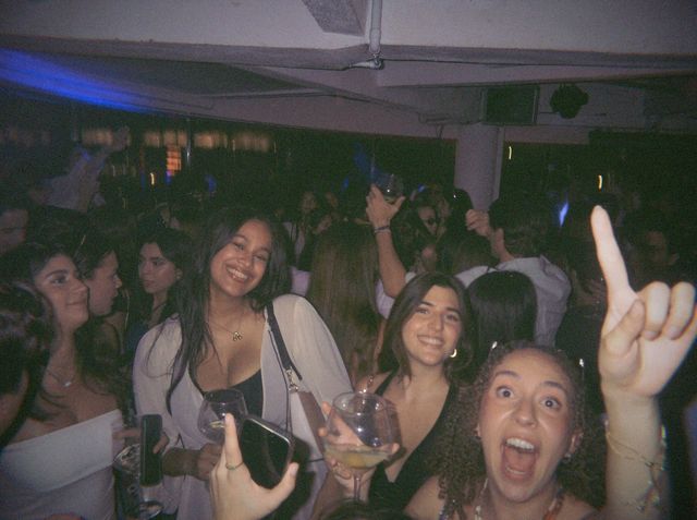 Una chica de pelo rizado grita con el índice en alto mientras sus amigas sonríen con copas en la pista.