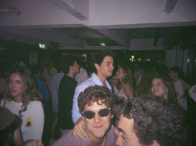 Un chico con gafas de sol redondas mira a cámara en la pista abarrotada, con la mano de alguien en el hombro.