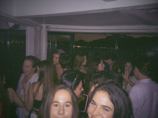 Dos chicas se asoman a cámara en primer plano entre la gente, con luces lejanas tras los ventanales.