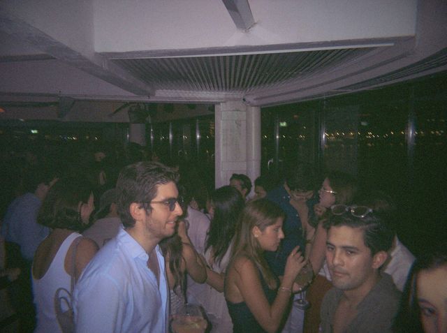 Un chico con gafas de sol y camisa blanca mira a un lado entre la gente; delante, otro lleva las gafas en la cabeza.