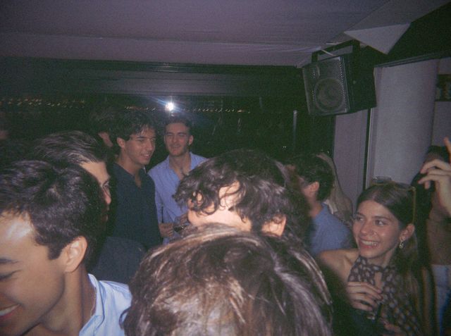 Una chica con gafas de sol en la cabeza ríe entre un grupo de chicos apretados bajo un altavoz.