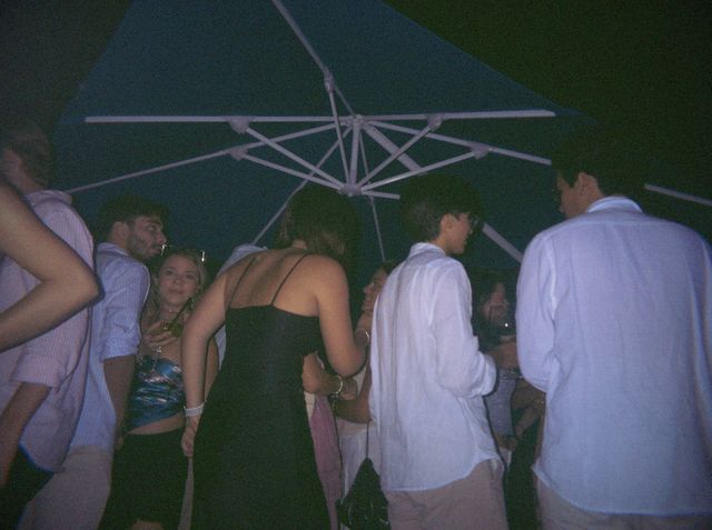 Una chica con top azul satinado mira a la cámara entre la gente reunida bajo una sombrilla azul en la terraza.
