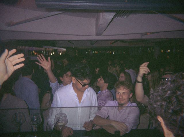 La gente baila con las manos en alto tras un cristal donde se apoyan un chico con gafas de sol y otro de camisa rosa.