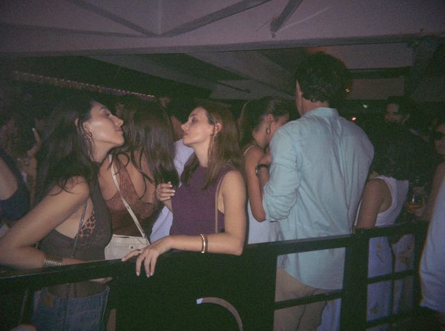 Dos chicas se miran casi nariz con nariz, apoyadas en una barandilla negra entre la gente.