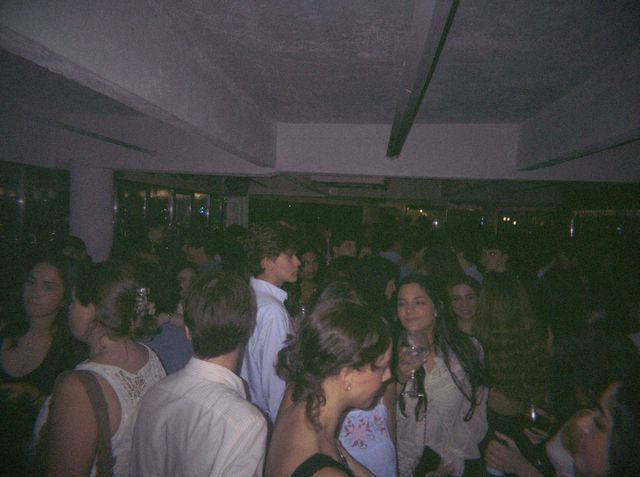 La gente llena la sala y charla, y una chica de pelo largo con blusa de encaje sostiene una copa.