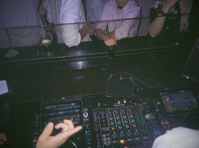 Una mano hace un gesto sobre la mesa de mezclas mientras la gente se apoya en la cabina del DJ.