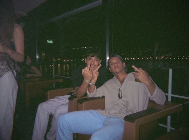 Dos chicos sentados en butacas junto al ventanal hacen la peineta a la cámara, con las luces de la bahía detrás.