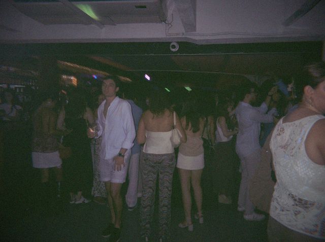 Un chico con camisa blanca abierta y una copa en la mano mira a cámara en la pista, entre gente vestida de blanco.