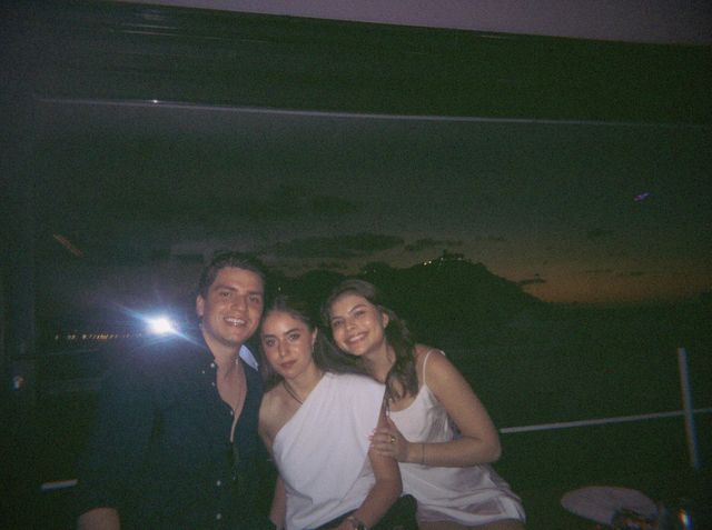 Un chico y dos chicas vestidas de blanco posan muy juntos ante el ventanal, con la silueta de un monte detrás.