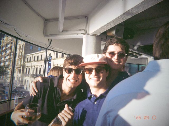 Tres amigos con gafas de sol se abrazan y sonríen junto a una ventana soleada; uno lleva gorra y otro, una copa.