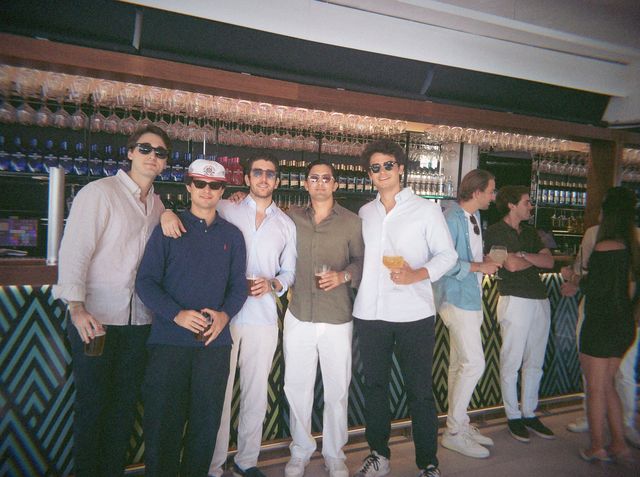 Cinco chicos con gafas de sol posan en la barra con sus bebidas; uno lleva una gorra blanca.